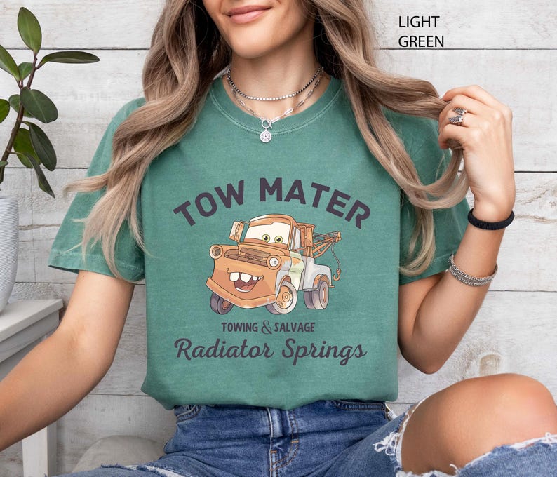 May include: Light green t-shirt featuring a cartoon image of Tow Mater from the movie Cars. The text "TOW MATER" arches above the image, with "Towing & Salvage" and "Radiator Springs" below. The shirt is a comfortable, casual style.