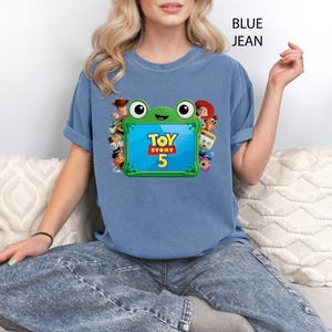 May include: A blue jean coloured t-shirt featuring a graphic of a green frog-shaped frame containing the text Toy Story 5. The frame is surrounded by various animated characters including a cowboy, a cowgirl, and other figures from the film series.
