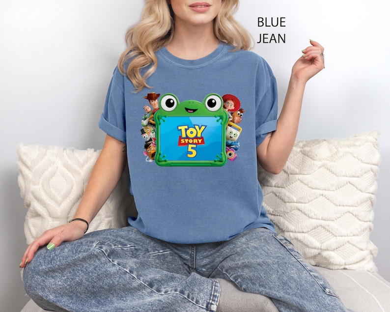 May include: A blue jean coloured t-shirt featuring a graphic of a green frog-shaped frame containing the text Toy Story 5. The frame is surrounded by various animated characters including a cowboy, a cowgirl, and other figures from the film series.
