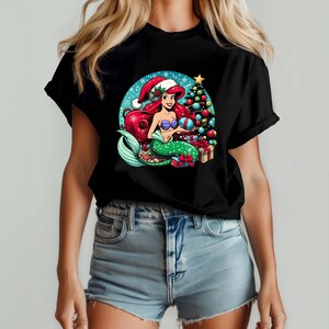 Puede incluir: Camiseta negra con un dise&ntilde;o navide&ntilde;o. Representa a una sirena pelirroja, con un gorro de Pap&aacute; Noel y un regalo. Un &aacute;rbol de Navidad y regalos tambi&eacute;n est&aacute;n en el dise&ntilde;o. La sirena tiene una cola verde y un sujetador de concha p&uacute;rpura.
