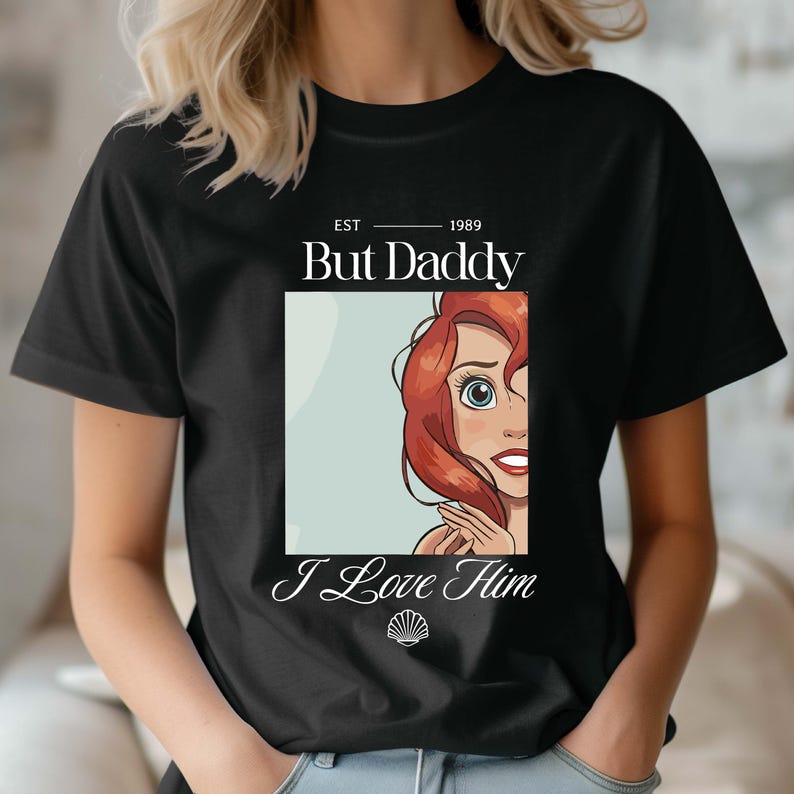 May include: Black t-shirt with the text "But Daddy" above an illustration of a cartoon character with red hair and blue eyes. The text "I Love Him" is below the illustration.