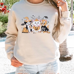 May include: Beige sweatshirt featuring a Halloween-themed graphic of three cartoon cats. The cats are dressed in costumes, including a witch hat, a pumpkin, and a skeleton outfit. The design includes a spider web and the word "RIP" on a tombstone.