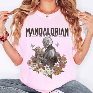 Puede incluir: Camiseta rosa claro con un dise&ntilde;o de The Mandalorian y Grogu, rodeados de flores y mariposas. El texto "MANDALORIAN" y "THIS IS THE WAY" est&aacute; impreso encima de la imagen. La camiseta es de color rosa claro.