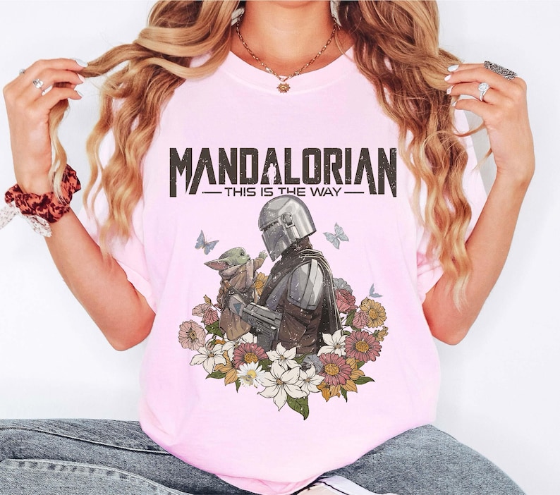 Puede incluir: Camiseta rosa claro con un dise&ntilde;o de The Mandalorian y Grogu, rodeados de flores y mariposas. El texto "MANDALORIAN" y "THIS IS THE WAY" est&aacute; impreso encima de la imagen. La camiseta es de color rosa claro.