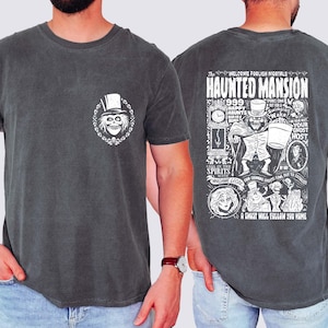 May include: Dark gray t-shirt featuring a white graphic of a skull wearing a top hat on the front. The back of the shirt displays a detailed illustration with the words "HAUNTED MANSION" and other text and images. The shirt is a crew neck.