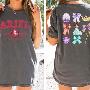 Może przedstawiać: Ciemnoszara koszulka z napisem "ARIEL" w czerwonych literach i czerwoną grafiką syreny. Tył koszulki przedstawia ilustracje korony, kokard, muszli i tr&oacute;jzębu. Widoczny jest r&oacute;wnież tekst "EST. 1989" i "SEA PRINCESS".