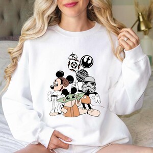 Puede incluir: Sudadera blanca con un dise&ntilde;o de Mickey Mouse, Baby Yoda y personajes de Star Wars. El dise&ntilde;o incluye a BB-8, un Stormtrooper y otros elementos ic&oacute;nicos de los universos de Star Wars y Disney. Cuello redondo.
