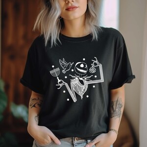 Op de afbeelding: Zwart t-shirt met een witte afbeelding van een skeletachtig personage in een gestreept pak, met een menorah, een duif en een tas. Het ontwerp bevat sterren en een rechthoekig kader.