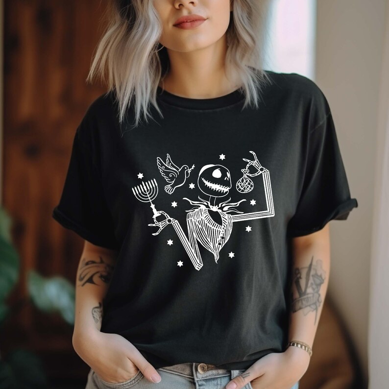 Op de afbeelding: Zwart t-shirt met een witte afbeelding van een skeletachtig personage in een gestreept pak, met een menorah, een duif en een tas. Het ontwerp bevat sterren en een rechthoekig kader.