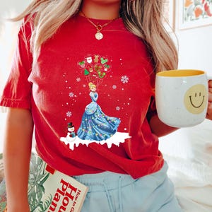 Op de afbeelding: Rood t-shirt met een afbeelding van een prinses in een blauwe jurk die kerstballonnen vasthoudt, met een sneeuwpop en sneeuwvlokken. Het woord "RED" staat bovenaan gedrukt. Het shirt wordt gedragen met een blauwe short.