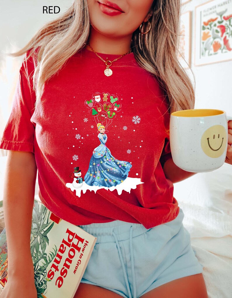 Op de afbeelding: Rood t-shirt met een afbeelding van een prinses in een blauwe jurk die kerstballonnen vasthoudt, met een sneeuwpop en sneeuwvlokken. Het woord "RED" staat bovenaan gedrukt. Het shirt wordt gedragen met een blauwe short.