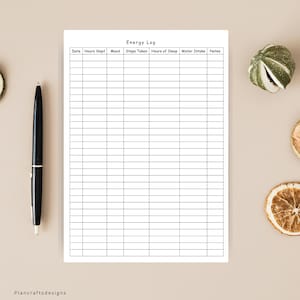 May include: A white paper energy log tracker with black grid lines and headers for date, hours slept, mood, steps taken, hours of sleep, water intake, and notes. A black ballpoint pen is positioned vertically to the left of the paper sheet.