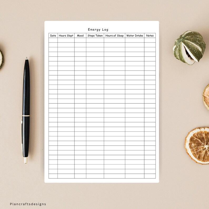 May include: A white paper energy log tracker with black grid lines and headers for date, hours slept, mood, steps taken, hours of sleep, water intake, and notes. A black ballpoint pen is positioned vertically to the left of the paper sheet.