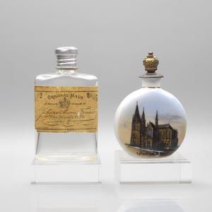 May include: A clear rectangular glass bottle with a silver cap and a tan label reading Original Haus Johann Maria Farina, alongside a round white porcelain bottle featuring a painted cathedral and a small gold crown-shaped stopper.