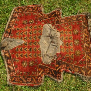 Könnte beinhalten: Ein antiker, rot-brauner Teppich mit floralem Muster und Fransen. Der Teppich hat ein abgenutztes Aussehen und liegt auf grünem Gras. Ein Leder-Sattelteil liegt oben auf dem Teppich.