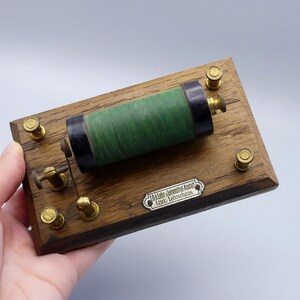 Può includere: Un componente elettrico antico con base in legno e hardware in ottone. Una bobina verde è montata sulla base in legno, con un cilindro nero ad ogni estremità. Una piccola placca metallica con testo è fissata alla base.