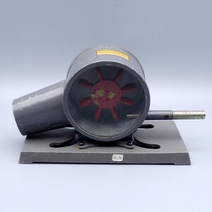 Könnte beinhalten: Ein grauer Ventilator im Vintage-Stil mit einem roten Lüfterrad in einem kreisförmigen Gehäuse. Der Ventilator ist auf einem grauen Sockel montiert, auf dem die Nummer "No. 34" zu sehen ist. Der Ventilator hat einen zylindrischen Körper und einen Griff.