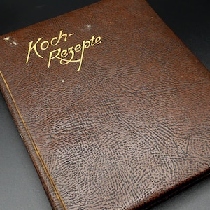 May include: A brown, textured cookbook with the gold text "Koch-Rezepte" on the cover. The book has a rectangular shape and appears to be vintage. The cover has a leather-like appearance.