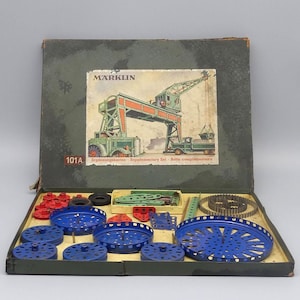 Puede incluir: Juego de construcción Märklin vintage en una caja verde. La tapa de la caja presenta una ilustración de una grúa. En el interior, varios componentes metálicos en rojo, verde y azul están dispuestos, incluyendo ruedas y engranajes.
