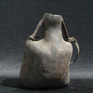 Pode incluir: Cantil de couro castanho escuro, de estilo antigo e com aspeto desgastado. Apresenta um corpo largo e arredondado e um pescoço estreito com uma pega de correia de couro. O cantil é provavelmente usado para transportar água ou outros líquidos.