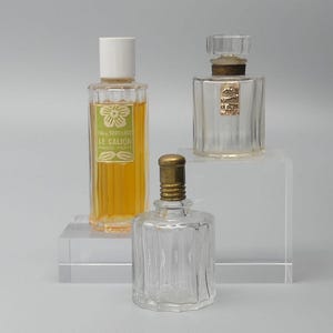 May include: Three vintage perfume bottles on a clear acrylic display. The bottles are clear glass with various shapes and stoppers. One bottle has a white cap and a label that reads "LE GALION". The perfume inside is amber-coloured.