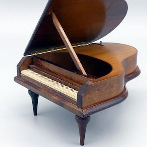 May include: A miniature wooden grand piano with a polished, dark brown finish. The piano's lid is open, revealing the keys and interior details. It has four legs, two are black and two are brown.