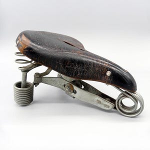 May include: Vintage bicycle seat with a distressed black leather top and metal springs. The seat has a classic design with a worn appearance, showcasing its age and use. The metal components are silver.