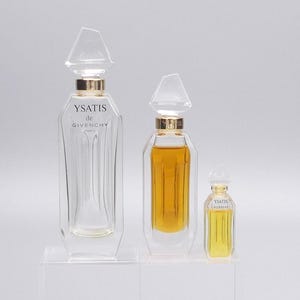 May include: Three clear glass perfume bottles with gold detailing and geometric stoppers. The bottles are labelled "YSATIS de GIVENCHY" and vary in size, with one containing a golden liquid. The bottles are displayed on clear stands.