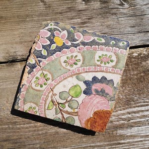 May include: A fragment of a decorative tile with a floral pattern. The tile displays pink, yellow, and blue flowers, green leaves, and a white backdrop. The tile is cracked and has a weathered look, set on a wooden surface.