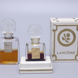 Könnte beinhalten: Vintage Lancôme Parfümflaschen und -box. Zwei transparente Glasparfümflaschen mit dekorativen Verschlüssen, eine mit der Aufschrift "Cuir" und die andere mit "Jeuxtidies". Eine weiße Schachtel mit goldenen Akzenten und dem Lancôme-Logo ist ebenfalls vorhanden.