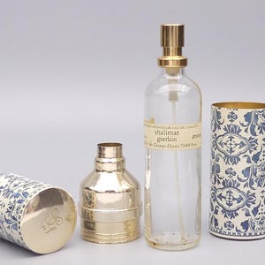 Può includere: Un assortimento di flaconi di profumo e confezioni Guerlain Shalimar. Il set include un flacone di vetro trasparente con un vaporizzatore dorato, un flacone in metallo color oro e due contenitori cilindrici con un motivo floreale blu e bianco. La confezione ha dettagli dorati.