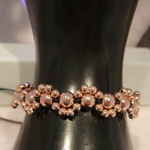 May include: A bracelet featuring a repeating pattern of metallic rose gold-colored spherical beads of varying sizes. The beads are arranged in a circular, flower-like cluster design and are strung together on a thin, dark black cord.