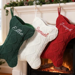 May include: Three cable-knit Christmas stockings hanging in a row. The left stocking is dark green with the name Ellie embroidered in white. The center stocking is white with the name Samuel in red. The right stocking is red with Joseph in white.