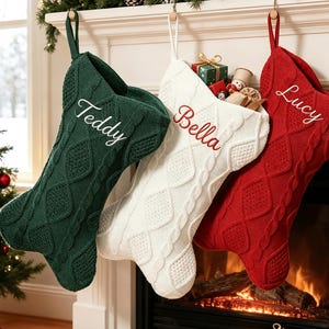 May include: Three cable-knit Christmas stockings in dark green, white, and red. The green stocking is embroidered with the name Teddy, the white with Bella, and the red with Lucy. Each stocking features a textured diamond pattern and a hanging loop.