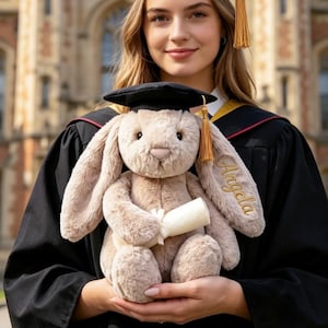 May include: A plush beige rabbit toy wearing a black graduation cap with a gold tassel, holding a small white rolled diploma. The rabbit has the name Angela embroidered in gold thread on one of its long, soft, floppy ears.