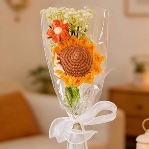 May include: A bouquet of crocheted flowers, including a large sunflower with a brown center and yellow petals, and an orange flower. The bouquet is wrapped in clear cellophane and tied with a white ribbon. The bouquet is in a clear vase.