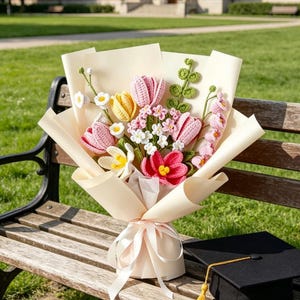 May include: A bouquet of crocheted flowers in shades of pink, yellow, red, and white, wrapped in cream-coloured paper and tied with a ribbon. A black graduation cap with a gold tassel rests on a wooden bench.