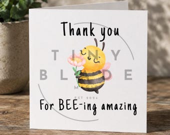 Può includere: Un biglietto di auguri bianco con la scritta "Thank you" in nero. Al centro c'è un'ape dei cartoni animati che tiene in mano un fiore rosa. Il testo "For BEE-ing amazing" è in basso. Il biglietto ha un design floreale e le parole "TINY BLADE" in un cerchio.