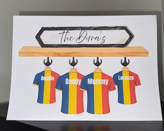 Può includere: Stampa incorniciata con una mensola in legno con quattro magliette appese sotto. Ogni maglietta ha una combinazione di colori diversa e un nome: Amalia, Daddy, Mummy e Lorenzo. Il testo "The Dura's" è sopra la mensola.