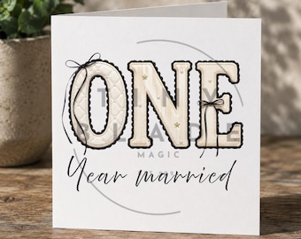 Può includere: Un biglietto bianco con la parola "ONE" in grandi lettere decorative. La "O" è trapuntata con un bordo e un fiocco neri. Le parole "Year married" sono scritte in corsivo sotto la parola "ONE".