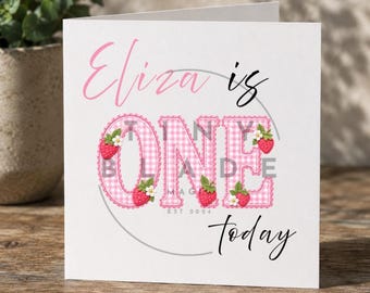 Può includere: Biglietto di auguri bianco con la scritta "Eliza is ONE today" in rosa e nero. La parola "ONE" è in un motivo a quadretti rosa, decorata con fragole e fiori. Il biglietto è per un primo compleanno.