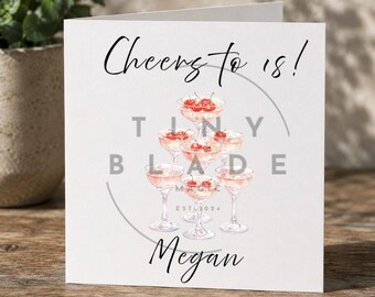 Può includere: Biglietto di auguri bianco con la scritta "Cheers to 18!" in corsivo nero. Il biglietto presenta un'illustrazione ad acquerello di calici di champagne impilati a piramide, ognuno sormontato da ciliegie. Il nome "Megan" è scritto sotto.