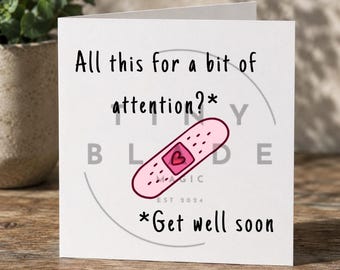 Può includere: Un biglietto di auguri bianco con il testo "All this for a bit of attention?*" e "*Get well soon". Un cerotto rosa con un cuore è al centro. Il biglietto è su una superficie di legno.