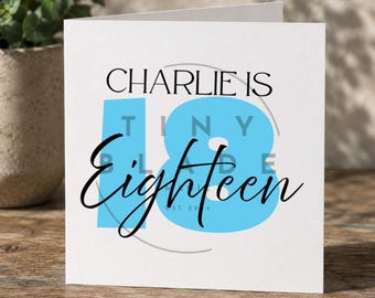 Può includere: Un biglietto di auguri bianco con la scritta "CHARLIE IS EIGHTEEN" in corsivo nero e una grande grafica "18" azzurra. La parola "TINY" è all'interno del numero 18. Il biglietto è su una superficie di legno.