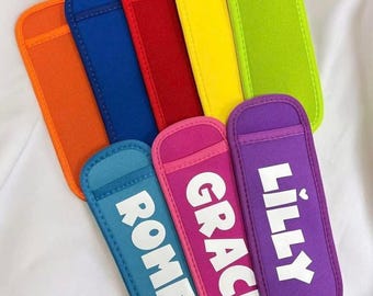 Può includere: Una collezione di colorate custodie termiche per bevande in varie tonalità, tra cui arancione, blu, rosso, giallo, verde, rosa e viola. Ogni custodia ha un nome bianco stampato: Romeo, Grace e Lilly. Le custodie sono progettate per mantenere fresche le bevande.