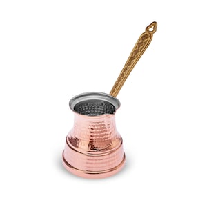 May include: A copper-colored Turkish coffee pot with a hammered texture. It has a silver-colored interior and a long, decorative wooden handle. The pot is designed for brewing coffee and has a traditional shape.