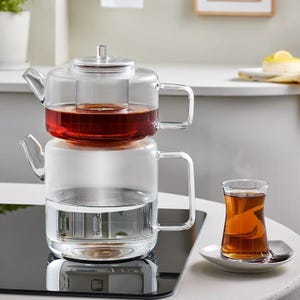 Puede incluir: Una tetera de vidrio transparente con infusor incorporado, llena de té rojo, se asienta sobre una base llena de agua. Un vaso de té en un platillo está al lado de la tetera. La tetera tiene un asa y una tapa.