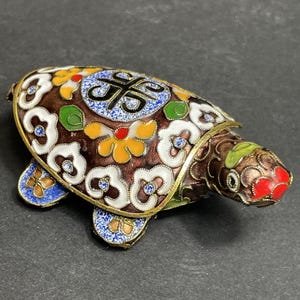 Può includere: Una statuetta decorativa a forma di tartaruga con un intricato lavoro di smalto cloisonné nei toni del marrone, bianco, giallo, rosso, verde e blu. Il guscio presenta un motivo circolare centrale con simboli neri circondati da motivi floreali.