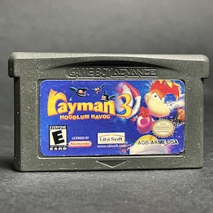 May include: A Game Boy Advance cartridge for the game "Rayman 3: Hoodlum Havoc." The label features the game's title in yellow and blue, with character illustrations. The cartridge is gray and includes the ESRB rating.