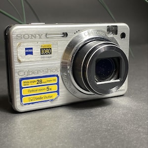 May include: A silver Sony Cyber-shot digital camera with a Carl Zeiss lens. The front features labels for Full HD 1080, a 28mm wide-angle lens, 5x optical zoom, and Smile Shutter technology. The camera body has a brushed metal finish and a black lens barrel.